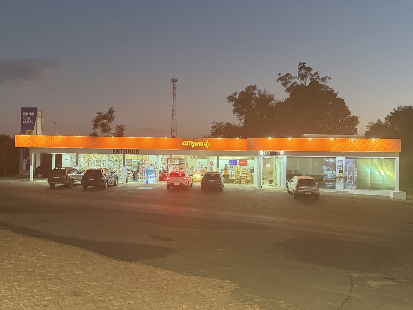 Fachada iluminada da loja de conveniência AmPm, com entrada e sanitários